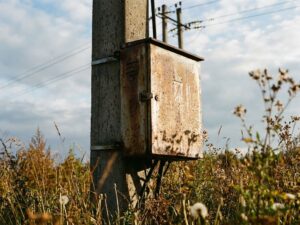 Verwitterter Stromverteilerkasten auf Betonmast mit Regentropfen, umgeben von Wildblumen und Gras
